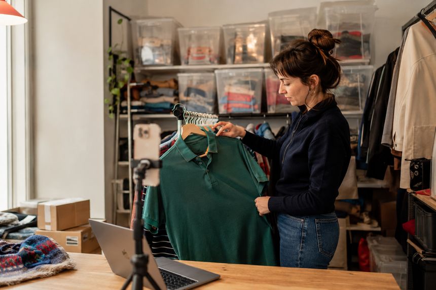 Une vendeuse présente un polo vert devant son portant, dans un atelier où des bacs transparents sont rangés sur des étagères. Un téléphone sur trépied et un ordinateur portable sont posés devant elle.